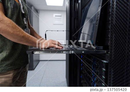 It technician working on a laptop console in a data center server rack performing network maintenance and monitoring It technician working on a laptop console in a data center server rack performing network maintenance and monitoring 137505420