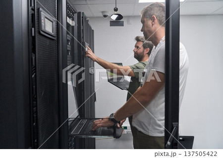 Two IT technicians troubleshooting servers in a secure data center server room with laptops Two IT technicians troubleshooting servers in a secure data center server room with laptops 137505422