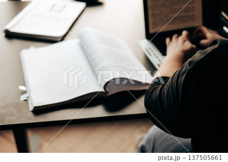 Unrecognizable person working on a laptop with an open book on a desk. Natural soft lighting, study or remote office atmosphere. Concept of education, research, and deep work. Lifestyle aesthetic. 137505661