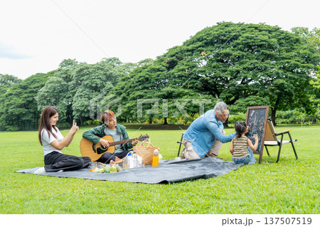 Asian young woman use mobile to take photo of senior man and little girl help to do drawing work together in public park with daylight. 137507519