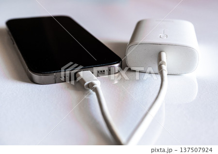 A smartphone is charging with a power bank. The phone is silver with a black screen. The power bank is white and connected with a charging cable. A smartphone is charging with a power bank. The phone is silver with a black screen. The power bank is white and connected with a charging cable. 137507924