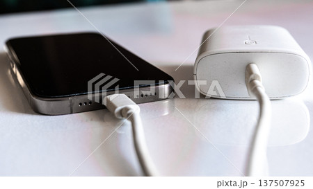 A smartphone is charging with a power bank. The phone is silver with a black screen. The power bank is white and connected with a charging cable. A smartphone is charging with a power bank. The phone is silver with a black screen. The power bank is white and connected with a charging cable. 137507925