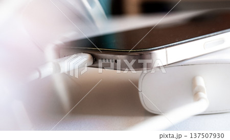 A smartphone is charging with a power bank. The phone is silver with a black screen. The power bank is white and connected with a charging cable. A smartphone is charging with a power bank. The phone is silver with a black screen. The power bank is white and connected with a charging cable. 137507930