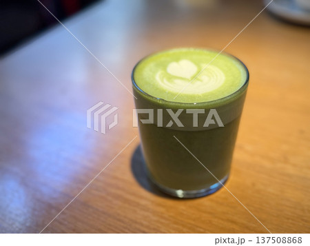Green matcha latte in glass cup with milk foam heart on wooden cafe table. Tea culture, calm ritual, modern beverage trend, mindfulness moment, relaxation and healthy lifestyle. Green matcha latte in glass cup with milk foam heart on wooden cafe table. Tea culture, calm ritual, modern beverage trend, mindfulness moment, relaxation and healthy lifestyle. 137508868