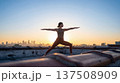 Woman practices yoga on rooftop sunrise. Warrior pose against city skyline, yoga inspiration for International Yoga Day, summer solstice wellness ads, copy space, mindful lifestyle concept 137508909