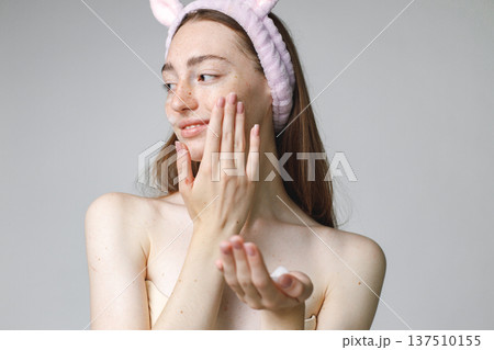 Close up beauty portrait of an attractive young woman apply a cream on her face over white background. Woman has no shirt. Woman has long red hair and wearing bunny ears make-up headband. Close up beauty portrait of an attractive young woman apply a cream on her face over white background. Woman has no shirt. Woman has long red hair and wearing bunny ears make-up headband. 137510155