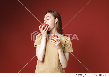 Studio portrait of caucasian girl isolated on red background. Portrait of young beautiful woman in beige t-shirt posing for a photo. Studio photo of a young girl holding two red apples. Studio portrait of caucasian girl isolated on red background. Portrait of young beautiful woman in beige t-shirt posing for a photo. Studio photo of a young girl holding two red apples. 137510174