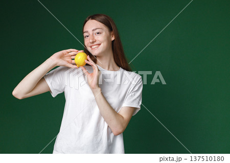Indoor portrait of caucasian girl isolated on green background. Portrait of young beautiful woman in white t-shirt posing for a photo. Studio photo of a young girl holding a lemon. Indoor portrait of caucasian girl isolated on green background. Portrait of young beautiful woman in white t-shirt posing for a photo. Studio photo of a young girl holding a lemon. 137510180