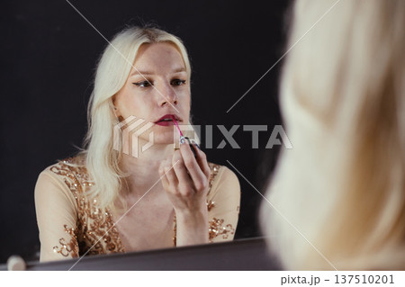 Young blond man holding brushes and doing make up on his face. Man wearing fashionable bright golden t-shirt. Man looking in a studio mirror. Young blond man holding brushes and doing make up on his face. Man wearing fashionable bright golden t-shirt. Man looking in a studio mirror. 137510201