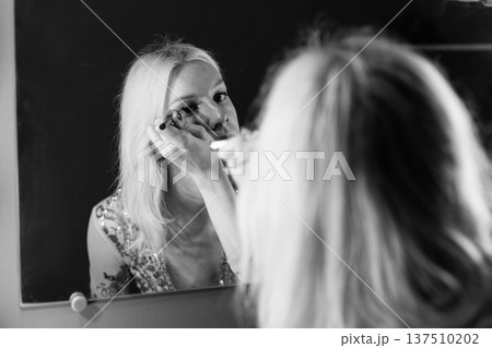 Young blond man holding brushes and doing make up on his face. Man wearing fashionable t-shirt. Man looking in a studio mirror. 137510202