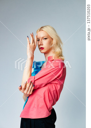 Young blond man has make up on one half of his face. Man wearing fashionable shirt divided on two colours bright pink and blue. Man isolated on grey background. 137510203