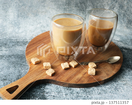 Two glasses of coffee with milk and sugar cubes on a wooden board Two glasses of coffee with milk and sugar cubes on a wooden board 137511357