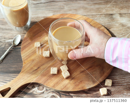 Hand Holding a Glass of Latte on a Wooden Board. Cappuccino or coffee drink with milk in tall glass cup Hand Holding a Glass of Latte on a Wooden Board. Cappuccino or coffee drink with milk in tall glass cup 137511360