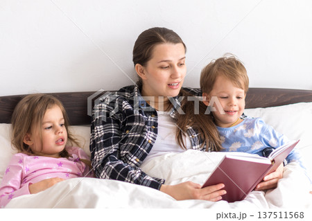 happy family. Close up loving mother lying with daughter son two kids pajamas in bed children reading interesting storybook, preschool smiling kid girl enjoy fairytale before go to sleep good pastime happy family. Close up loving mother lying with daughter son two kids pajamas in bed children reading interesting storybook, preschool smiling kid girl enjoy fairytale before go to sleep good pastime 137511568