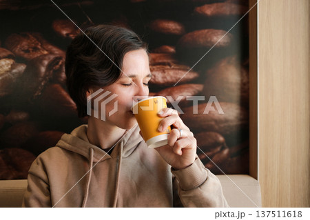 Young woman drinking coffee from a takeaway paper cup while sitting in a cafe with coffee bean background. Concept of coffee break and urban lifestyle Young woman drinking coffee from a takeaway paper cup while sitting in a cafe with coffee bean background. Concept of coffee break and urban lifestyle 137511618