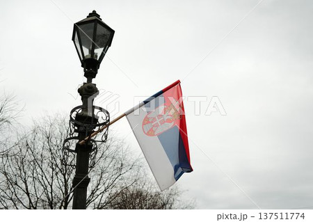 Serbian flag attached to vintage street lamp in Sombor, Serbia under cloudy sky. National identity and European urban atmosphere concept Serbian flag attached to vintage street lamp in Sombor, Serbia under cloudy sky. National identity and European urban atmosphere concept 137511774