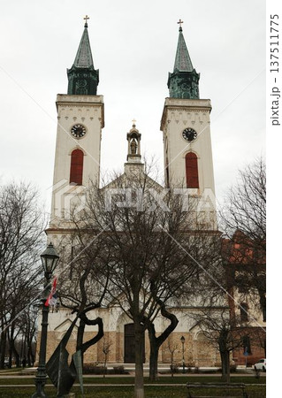 Twin church towers with clocks rising above winter trees in Sombor, Serbia. Symmetrical European landmark and cultural heritage concept Twin church towers with clocks rising above winter trees in Sombor, Serbia. Symmetrical European landmark and cultural heritage concept 137511775