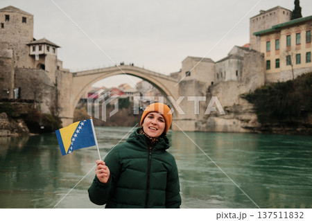A smiling woman holds the Bosnia and Herzegovina flag in front of the historic stone bridge by the river. Concept of travel memories, national identity, tourism lifestyle, and cultural exploration A smiling woman holds the Bosnia and Herzegovina flag in front of the historic stone bridge by the river. Concept of travel memories, national identity, tourism lifestyle, and cultural exploration 137511832