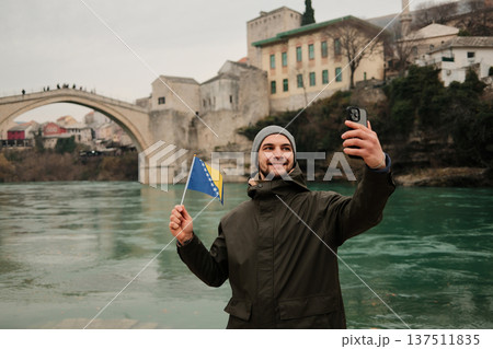 Happy traveler taking a selfie while holding the Bosnia flag near a famous old bridge in Mostar and Neretva river. Concept of tourism personal travel memory and Balkan culture Happy traveler taking a selfie while holding the Bosnia flag near a famous old bridge in Mostar and Neretva river. Concept of tourism personal travel memory and Balkan culture 137511835