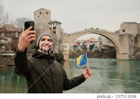 Happy traveler taking a selfie while holding the Bosnia flag near a famous old bridge in Mostar and Neretva river. Concept of tourism personal travel memory and Balkan culture Happy traveler taking a selfie while holding the Bosnia flag near a famous old bridge in Mostar and Neretva river. Concept of tourism personal travel memory and Balkan culture 137511836