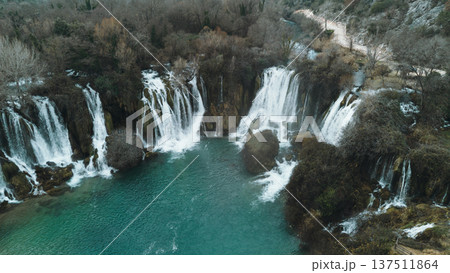 Aerial horizontal view of Kravica Waterfalls in Bosnia during winter, with powerful cascades flowing into a turquoise pool, surrounded by bare trees and rocky cliffs in a serene natural landscape Aerial horizontal view of Kravica Waterfalls in Bosnia during winter, with powerful cascades flowing into a turquoise pool, surrounded by bare trees and rocky cliffs in a serene natural landscape 137511864