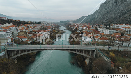 Aerial view of a road bridge crossing the Neretva River in Mostar, surrounded by residential houses with red tiled roofs, urban neighborhoods and hills under a cloudy sky Aerial view of a road bridge crossing the Neretva River in Mostar, surrounded by residential houses with red tiled roofs, urban neighborhoods and hills under a cloudy sky 137511878