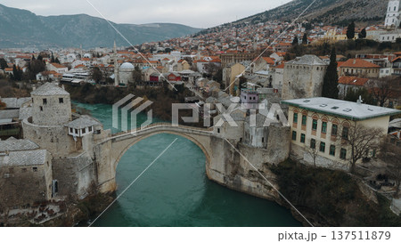 Aerial view of the historic stone bridge over the Neretva River in Mostar, with old town architecture, turquoise water, and surrounding mountains under an overcast sky. Amazing Bosnia and Herzegovina Aerial view of the historic stone bridge over the Neretva River in Mostar, with old town architecture, turquoise water, and surrounding mountains under an overcast sky. Amazing Bosnia and Herzegovina 137511879