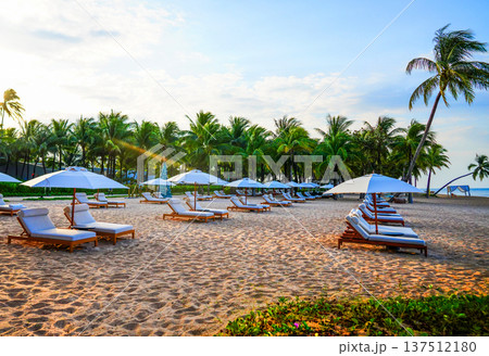 Tropical sand beach with palm trees on bright blue sky. Visitors relaxing on lounge chairs at sea. Tropical sand beach with palm trees on bright blue sky. Visitors relaxing on lounge chairs at sea. 137512180