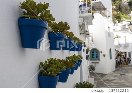 Beautiful blue pots with plants line the white walls of Frigiliana in Spain as people stroll through the streets during the day 137512216