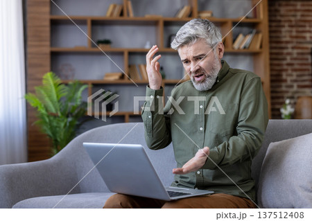 Mature man with gray hair sits on a sofa, frustrated and worried while working on a laptop at home, struggling with computer problems and connectivity issues Mature man with gray hair sits on a sofa, frustrated and worried while working on a laptop at home, struggling with computer problems and connectivity issues 137512408