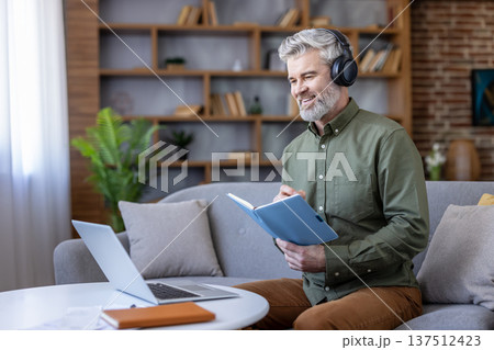 Mature man with headphones sitting on a sofa, looking at a laptop screen, taking notes in a notebook, concentrating on a virtual meeting or online course from his home office Mature man with headphones sitting on a sofa, looking at a laptop screen, taking notes in a notebook, concentrating on a virtual meeting or online course from his home office 137512423