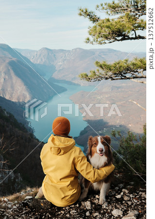 A woman sits at Banjska Stena viewpoint in Tara National Park holding an Australian Shepherd above the Drina River canyon. Concept of hiking travel, freedom, and deep bonding with pets in wild nature A woman sits at Banjska Stena viewpoint in Tara National Park holding an Australian Shepherd above the Drina River canyon. Concept of hiking travel, freedom, and deep bonding with pets in wild nature 137512662