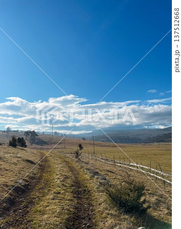 A dirt road crosses open fields and gentle hills in rural Bosnia under a wide blue sky. The scene represents freedom, travel, simplicity, and quiet countryside life A dirt road crosses open fields and gentle hills in rural Bosnia under a wide blue sky. The scene represents freedom, travel, simplicity, and quiet countryside life 137512668