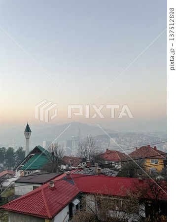 City view of Sarajevo with residential rooftops, mosque minaret, and distant urban skyline under soft evening haze. Calm Balkan atmosphere with layered cityscape 137512898