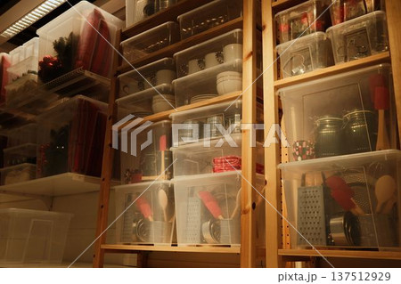 Wooden shelving with transparent plastic containers holding kitchen items, textiles, and decor in a storage room with warm indoor lighting 137512929