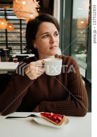 A woman holds a cup of cappuccino near a window with a strawberry tart on the table in a modern cafe interior 137512935