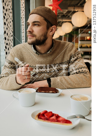 A man sits at a cafe table with cappuccino and desserts, holding a fork and looking away from the window in a cozy modern interior A man sits at a cafe table with cappuccino and desserts, holding a fork and looking away from the window in a cozy modern interior 137512938