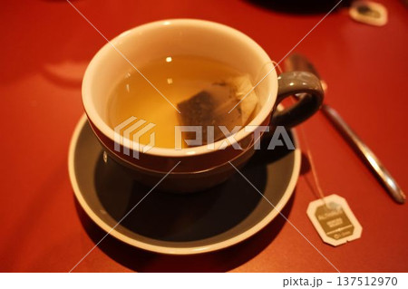 Hot tea in a ceramic cup with teabag resting inside, placed on a red table. Warm indoor light creates a cozy atmosphere of relaxation and quiet evening comfort Hot tea in a ceramic cup with teabag resting inside, placed on a red table. Warm indoor light creates a cozy atmosphere of relaxation and quiet evening comfort 137512970