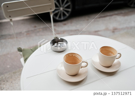 Two empty coffee cups and a metal ashtray with cigarette butts on a white outdoor table. Urban cafe atmosphere showing everyday habits, rest, and social routine Two empty coffee cups and a metal ashtray with cigarette butts on a white outdoor table. Urban cafe atmosphere showing everyday habits, rest, and social routine 137512971