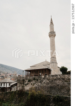 Mosque with tall minaret in Mostar old town surrounded by stone buildings and hillside landscape. Religious and cultural landmark of Balkan Islamic heritage 137512987