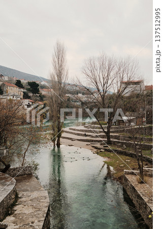Calm section of the Neretva River in Mostar with trees, stone embankments, and city houses along the water. Peaceful urban landscape combining nature and historic surroundings Calm section of the Neretva River in Mostar with trees, stone embankments, and city houses along the water. Peaceful urban landscape combining nature and historic surroundings 137512995