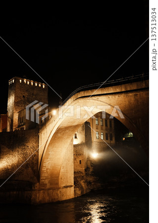 The iconic stone bridge of Mostar glows under night lighting above the river. Historic architecture and dramatic contrast highlight the landmark against the dark sky The iconic stone bridge of Mostar glows under night lighting above the river. Historic architecture and dramatic contrast highlight the landmark against the dark sky 137513034