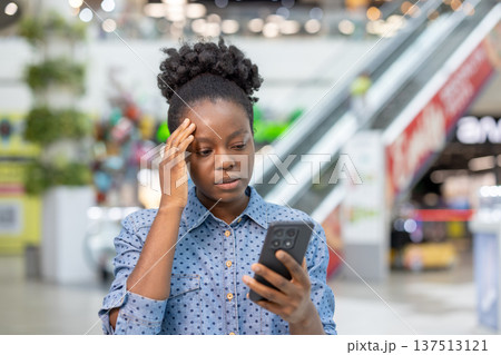 Young woman feeling upset and stressed while receiving troubling news on her mobile phone, experiencing a headache and worry inside a bustling modern shopping mall 137513121