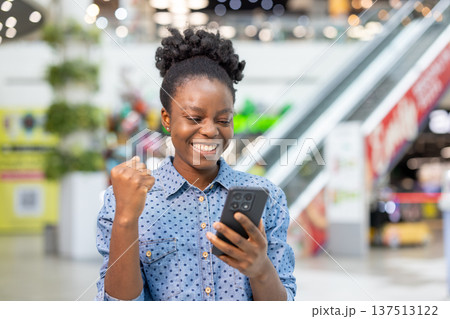 Happy young woman in a busy shopping mall celebrates good news on her smartphone, smiling and pumping a fist in triumph while browsing stores and using a mobile app 137513122
