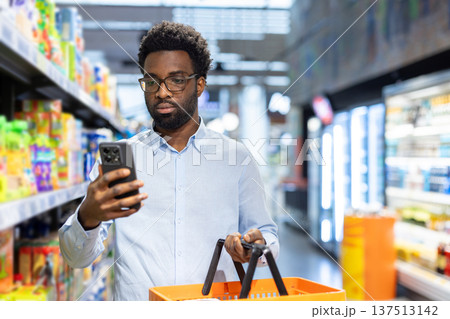 Young adult man standing in a grocery store aisle, comparing product information on his mobile phone while holding an orange shopping basket, engaging with modern retail technology 137513142