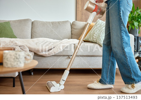 Woman vacuuming floor with cordless vacuum cleaner in home interior near sofa 137513493