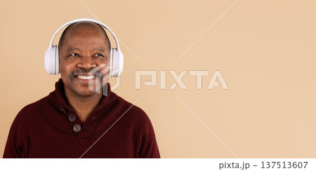 Smiling black man wearing white headphones listening to music on beige background Smiling black man wearing white headphones listening to music on beige background 137513607