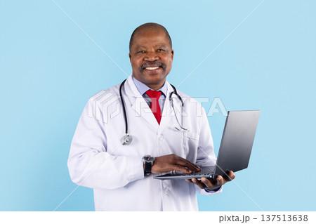 Smiling mature african american doctor using laptop computer on blue studio background Smiling mature african american doctor using laptop computer on blue studio background 137513638