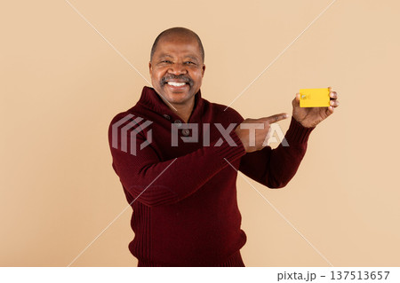 Mature black man pointing to credit card while smiling on beige background Mature black man pointing to credit card while smiling on beige background 137513657