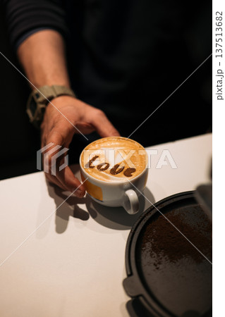 A hand holds a cup of latte art with the word 'Love' on top. The setting is a coffee shop with a blurred background. A hand holds a cup of latte art with the word 'Love' on top. The setting is a coffee shop with a blurred background. 137513682
